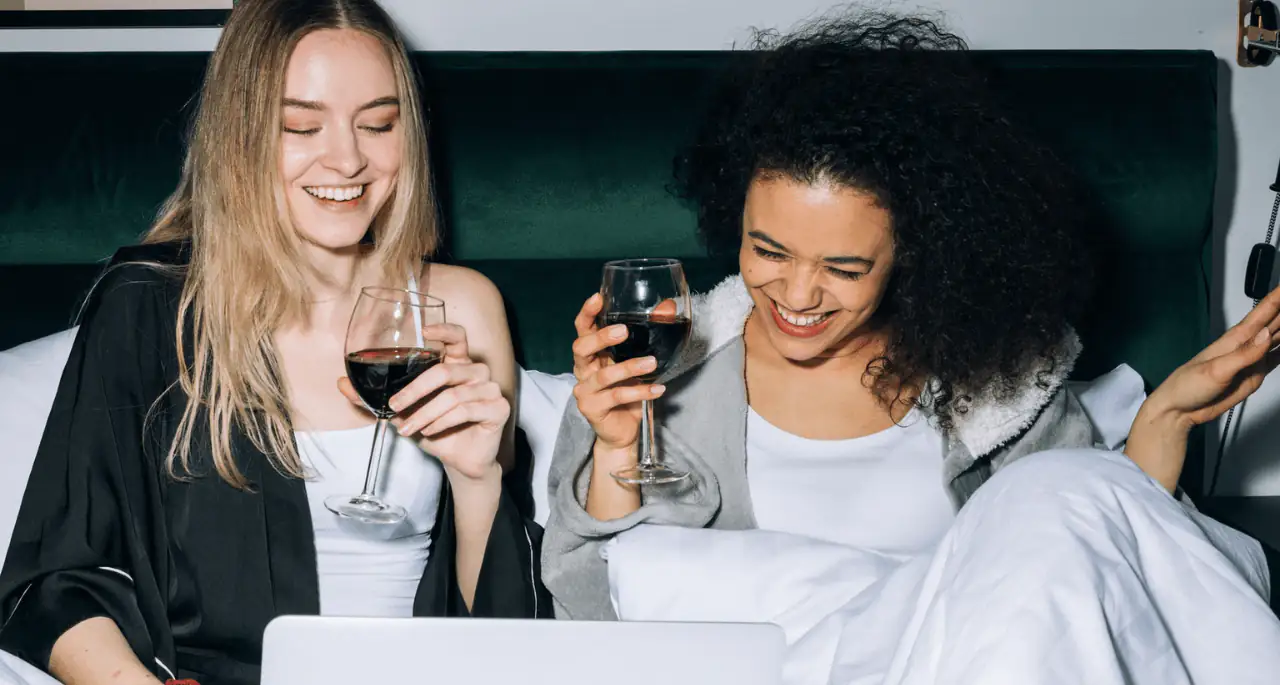 Two women drinking red wine in bed and laughing at something on a laptop Two women drinking red wine in bed and laughing at something on a laptop